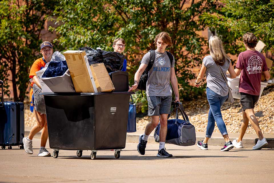 Students walk to their dorms carry items for their room, while another student pushes a cart filled to the brim with furniture.
