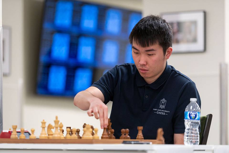 A student sitting a table with a chess board and a water bottle holds a chess piece while examining the board.