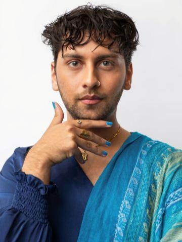 Anuj Gandhi poses for a photo against a white background. Anuj rests his right hand under his chin. Anuj wears a blue and teal multi-patterned long-sleeve shirt.