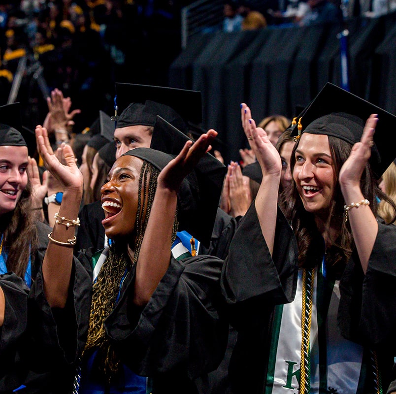 Two students in graduation gear smile and clap.