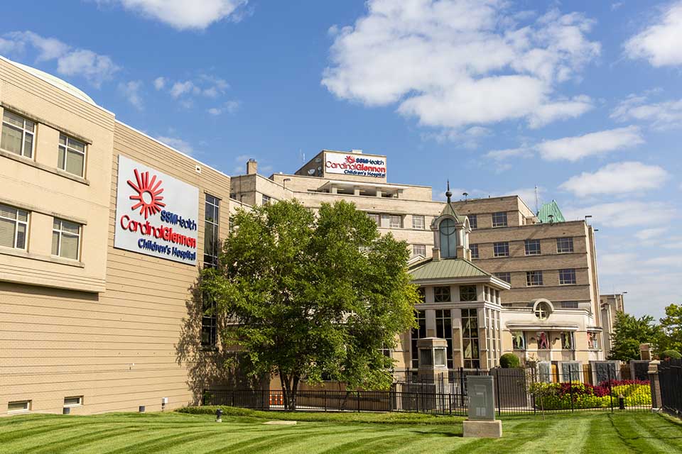 Sunny view of a historic children’s hospital with a red-and-blue SSM Health Cardinal Glennon sign, leafy tree, manicured lawn, and bright blue sky.