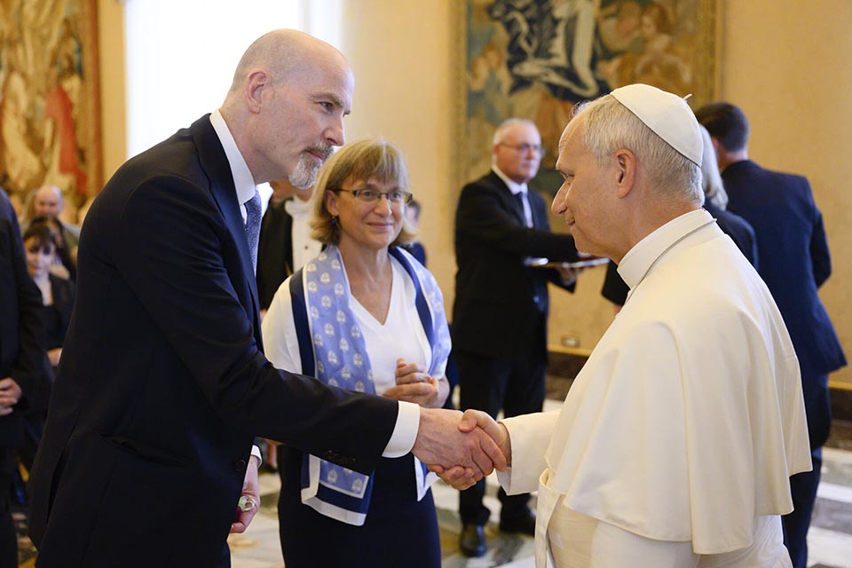 In an ornate room at the Apostolic Palace in Rome, SLU President Edward Feser, wearing a dark suit, shakes hands with Pope Leo XIV, wearing a white papal floor-length robe and cap, as Kathy Feaser looks on, wearing a white top and blue scarf.