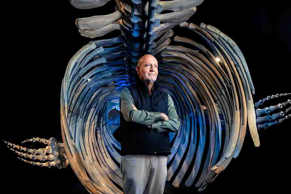 Jean Potvin stands confidently with his arms crossed in front of a large skeletal whale ribcage, dramatically lit in blue, conveying a sense of wonder and exploration.