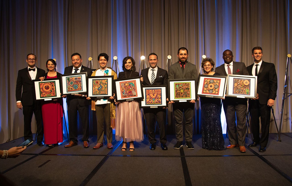Esmeralda Aharon (third from the right) poses with other awardees at the 2022 Adelante Awards. The photo is taken indoors in a dimly-lit room. The group stands on a stage holding framed artwork.