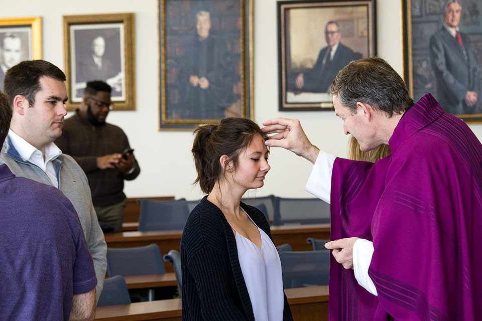 A female student with dark hair receives ashes on her forehead from a priest in a purple alb at an Ash Wednesday Mass celebrated at the School of Law's Scott Hall at Saint Louis University.