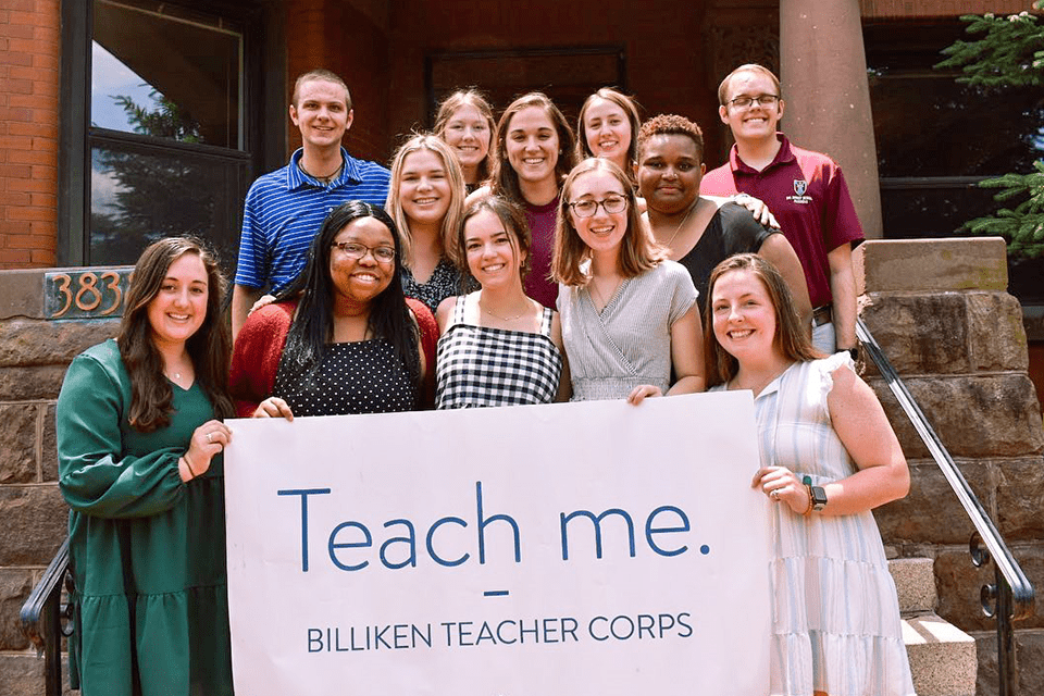 A group of SLU students hold a banner that reads "Teach Me. Billiken Teacher Corps" in front of a red brick building on SLU's campus. 