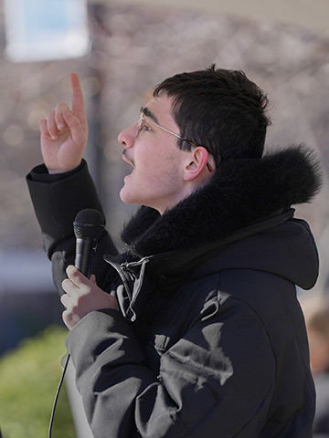 A photo student Nikolay Remizov wearing a winter coat and holding a microphone while speaking at the Campus Rally for Iran.