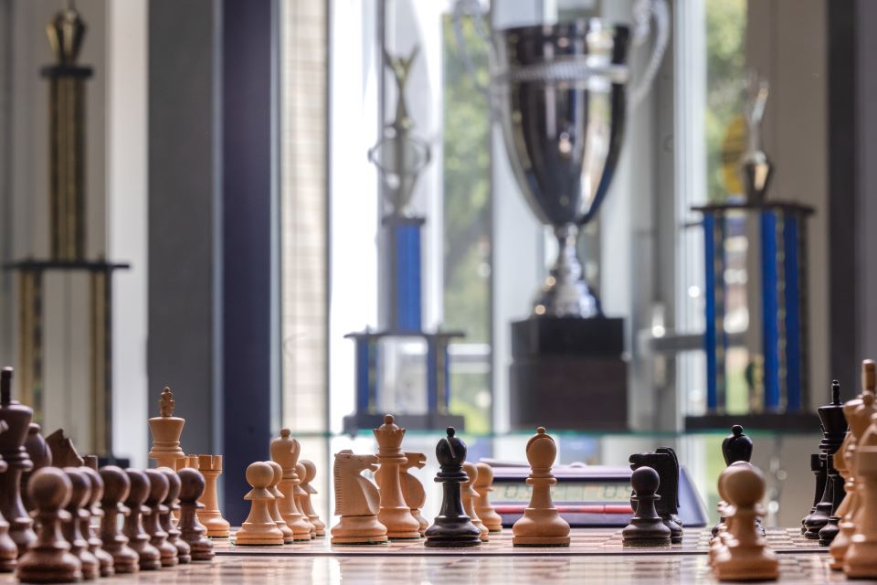 A closeup of a chess board on a brown table. Trophies in the background in front of a window.