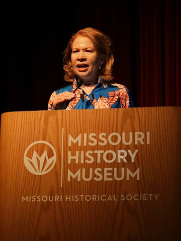 Angie Clay Thomas wears a blue and orange floral outfit as she speaks behind a podium labeled Missouri History Museum against a deep red curtain. Angie Clay Thomas wears a blue and orange floral outfit as she speaks behind a podium labeled Missouri History Museum against a deep red curtain.