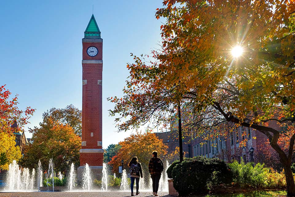 Two students walking through the Lipic Clock Tower Plaza and fountains with the sun shining through the fall leaves of a tree.