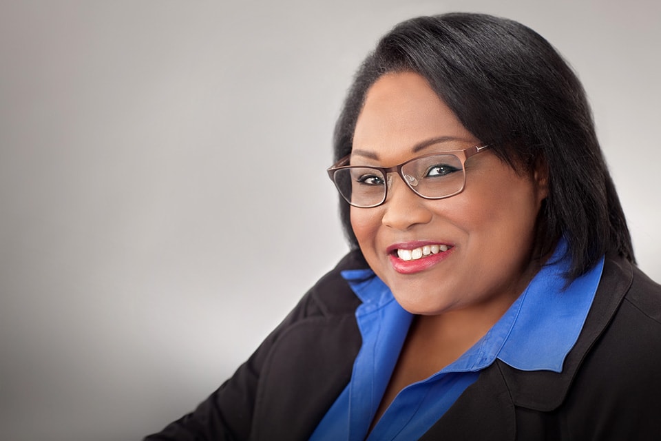 A headshot of a Black woman smiling, wearing a black suit with a blue shirt and black glasses. 