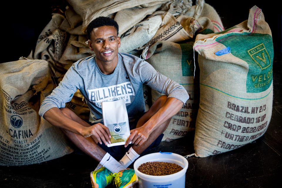 A student wearing a Billikens shirt holds a package of coffee, while sitting next to a bucket full of coffee beans. Several burlap sacks of coffee beans are in the background.