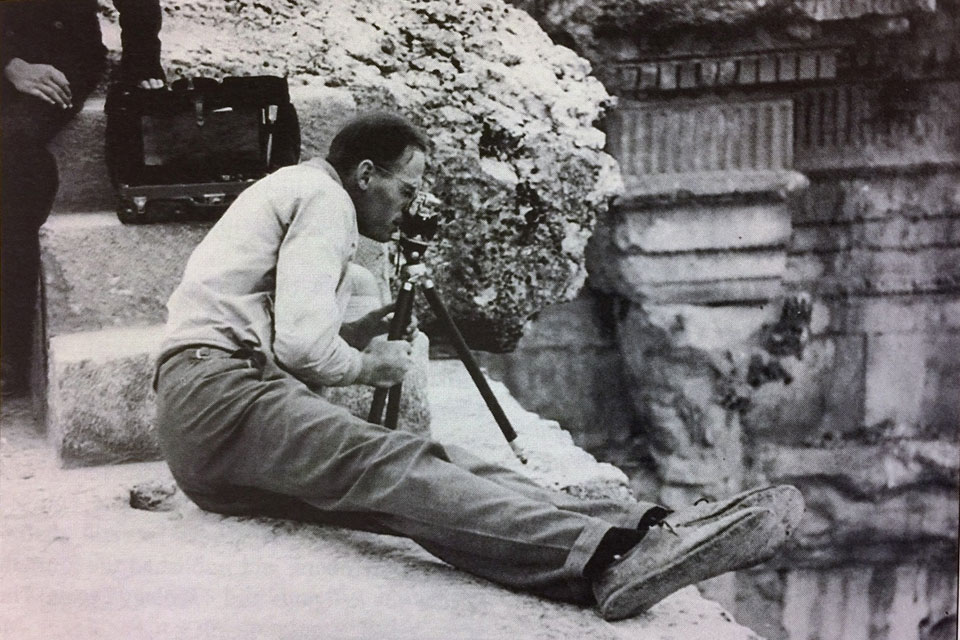 Claude Heithaus, S.J., surveys an archaeological site in the Middle East from a seated position with ruins in the background in an archival black and white photo.