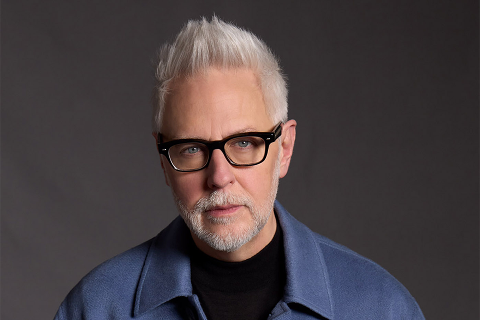 James Gunn, with short, spiky white hair and glasses, is wearing a blue collared jacket over a black shirt, set against a dark background.