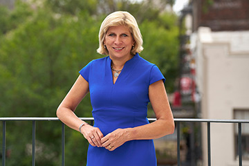 Dr. Kathy Johnson, with blond hair and wearing a royal blue dress, stands confidently on a balcony, smiling. Green trees and a blurred building form the background.