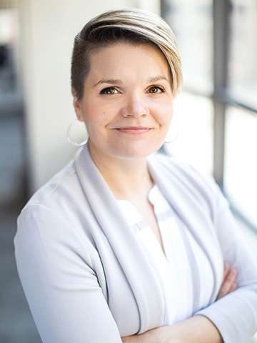 A woman with short blonde hair dressed in a light gray cardigan, smiling softly while standing against a neutral background