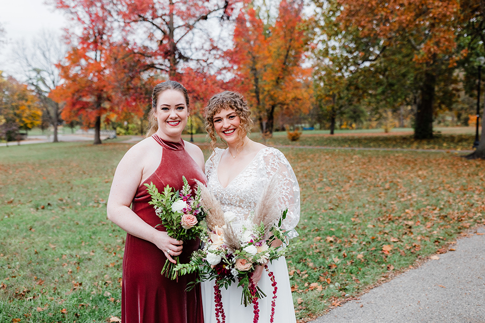 Two women dressed in wedding attire pose for a photo outdoors in a park on a fall day.