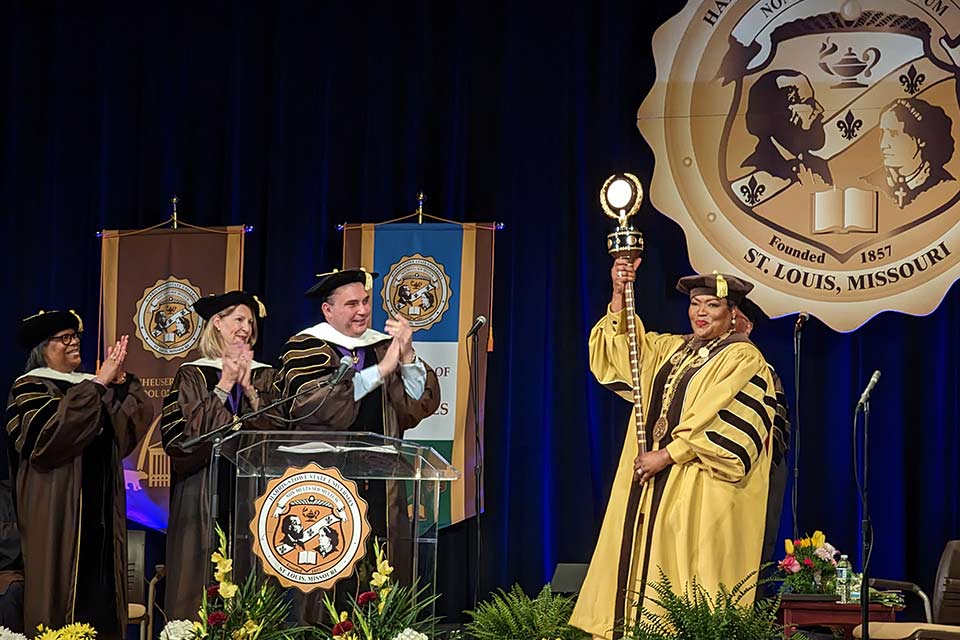 Dignitaries applaud for Harris-Stowe President LaTonia Collins Smith, who is wearing an academic cap and gown while holding HSSU’s Mace, a long wooden staff topped with a gold university seal and laurel wreath.