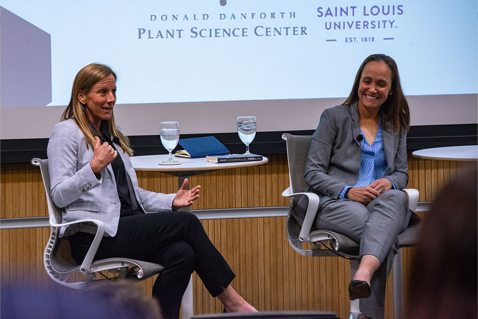 Two women are sitting, smiling and laughing while sitting in an auditorium discussing leadership for an audience. 