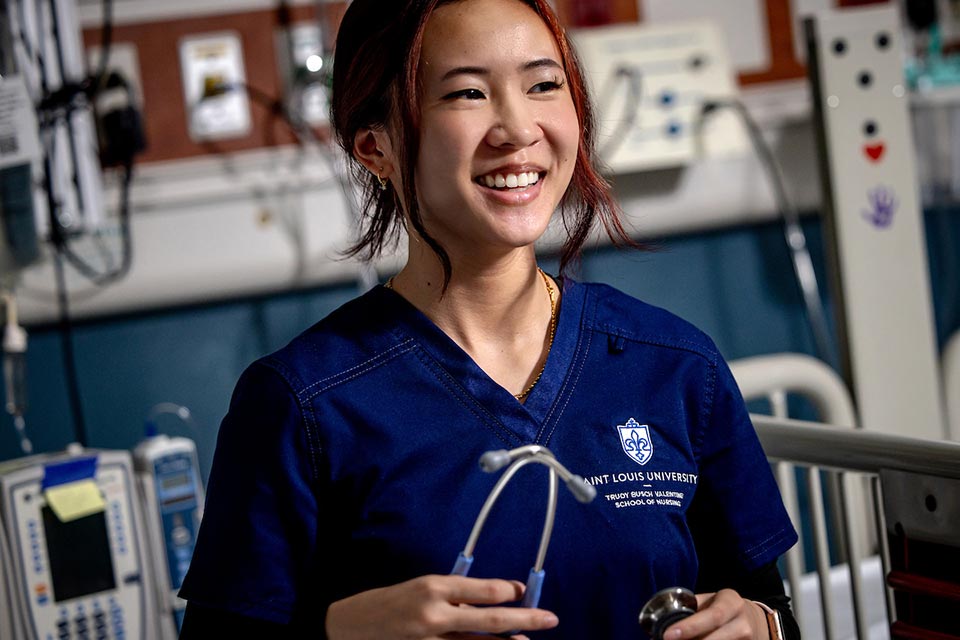 A nursing student smiles while wearing blue Saint Louis University scrubs and holding a stethoscope in a simulation training room. A nursing student smiles while wearing blue Saint Louis University scrubs and holding a stethoscope in a simulation training room.