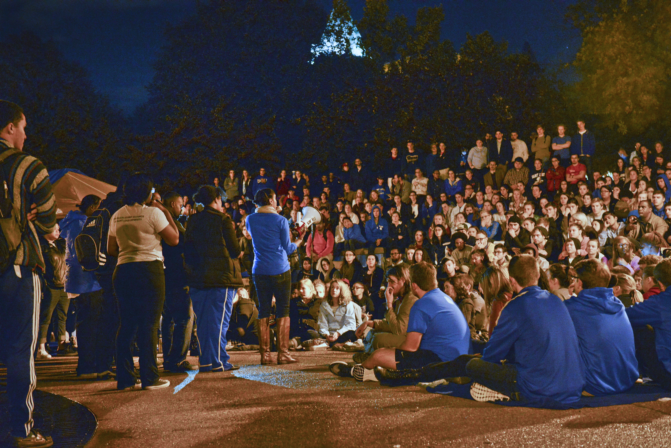 Students crowd around the clock tower plaza while listening to a speaker on a microphone