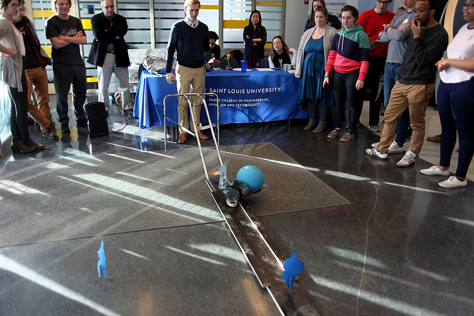 A competitor rolls his best Indiana Jones-style boulder (blue kick ball) down a ramp at the Parks College E-Week Innovation Challenge before a crowd.