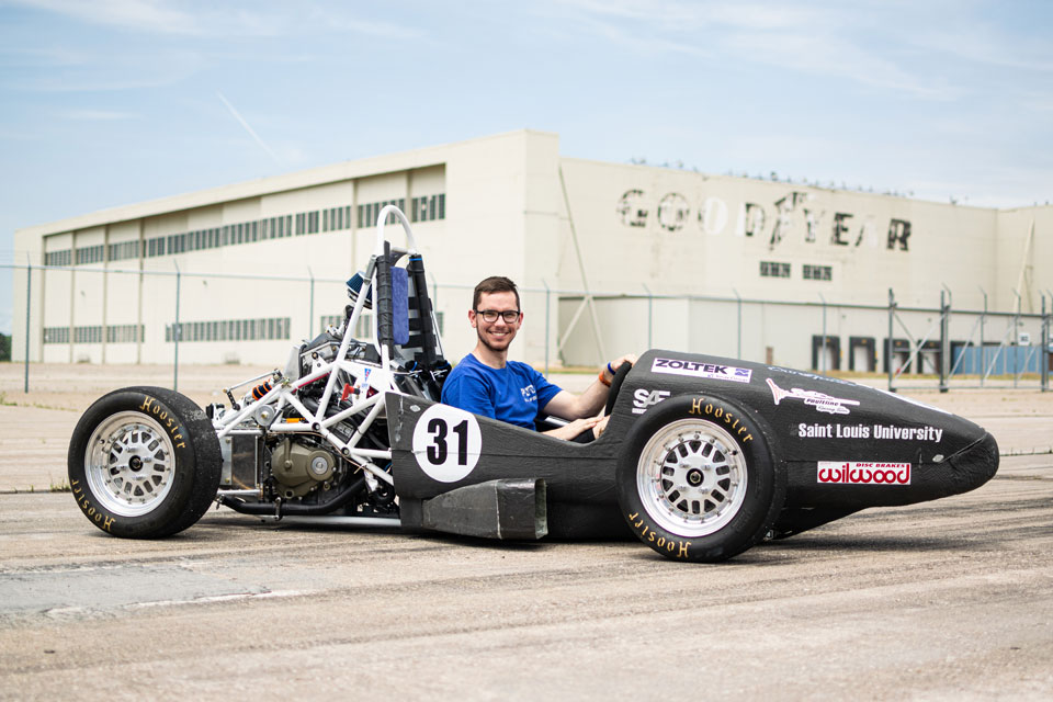 Jacob Bullard, a SLU student, poses in the race car he and other members of the Parks Racing FSAE Club built for competition. The car is black. Bullard has glasses, dark hair and a wide smile. He is seated in the car's driver's seat as it sits parked on a racetrack.