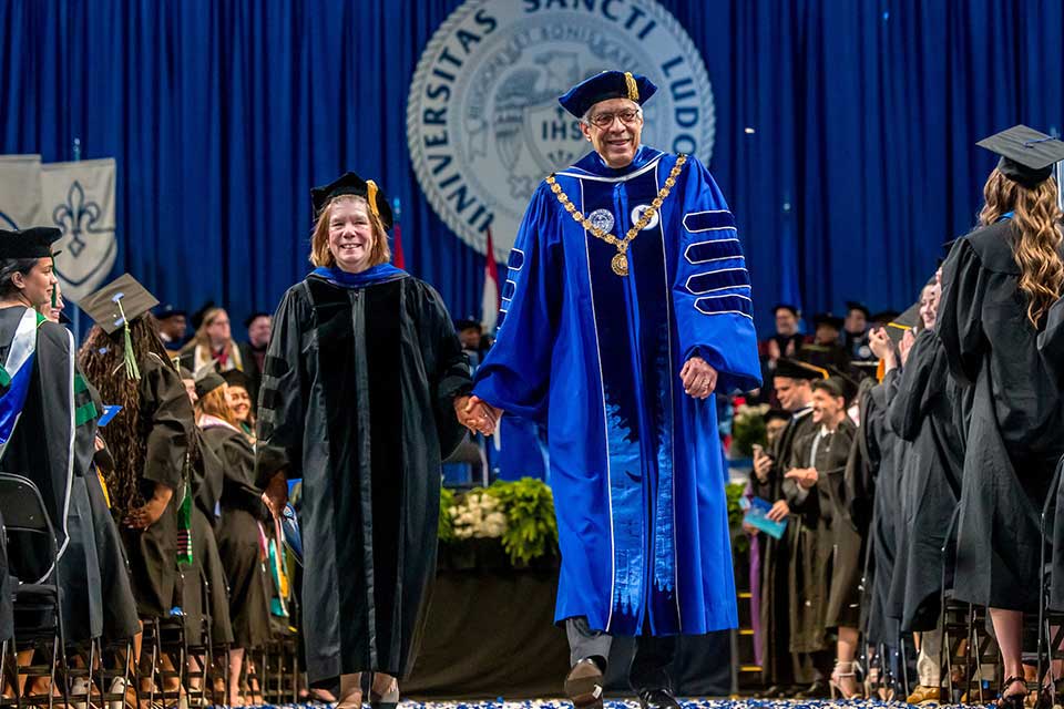 A woman and a man in academic regalia hold hands as they walk down an aisle with students in graduation attire on either side. 