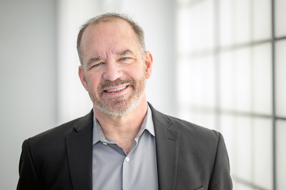 Professor Richard Grucza, Ph.D., wearing a black blazer and gray shirt, smiles warmly in a well-lit room with frosted windows, conveying a professional and friendly demeanor.