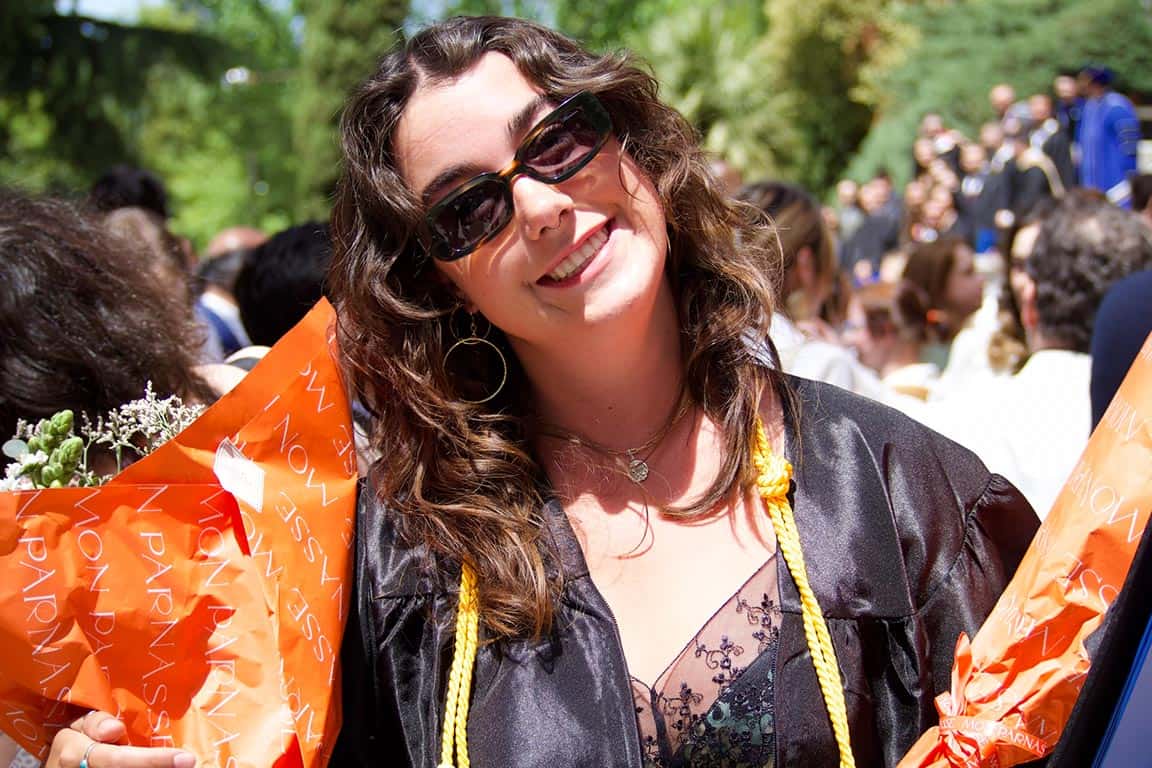 Quinn Riordan smiles for the camera. She wears a black graduation gown with a yellow tassel. She holds a bouquet of flowers in orange wrapping. 