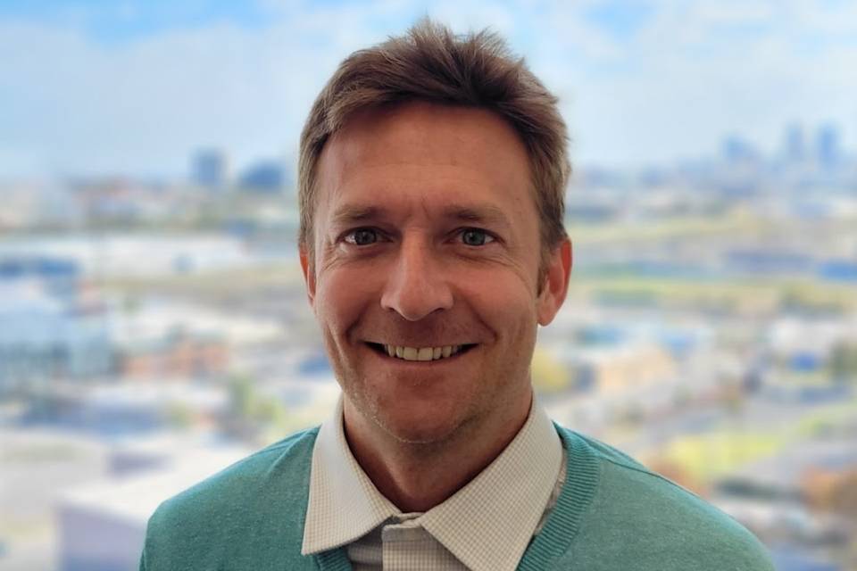 Professor Stephen Ferris smiles while wearing a teal green sweater, with a blurred cityscape visible in the background. 