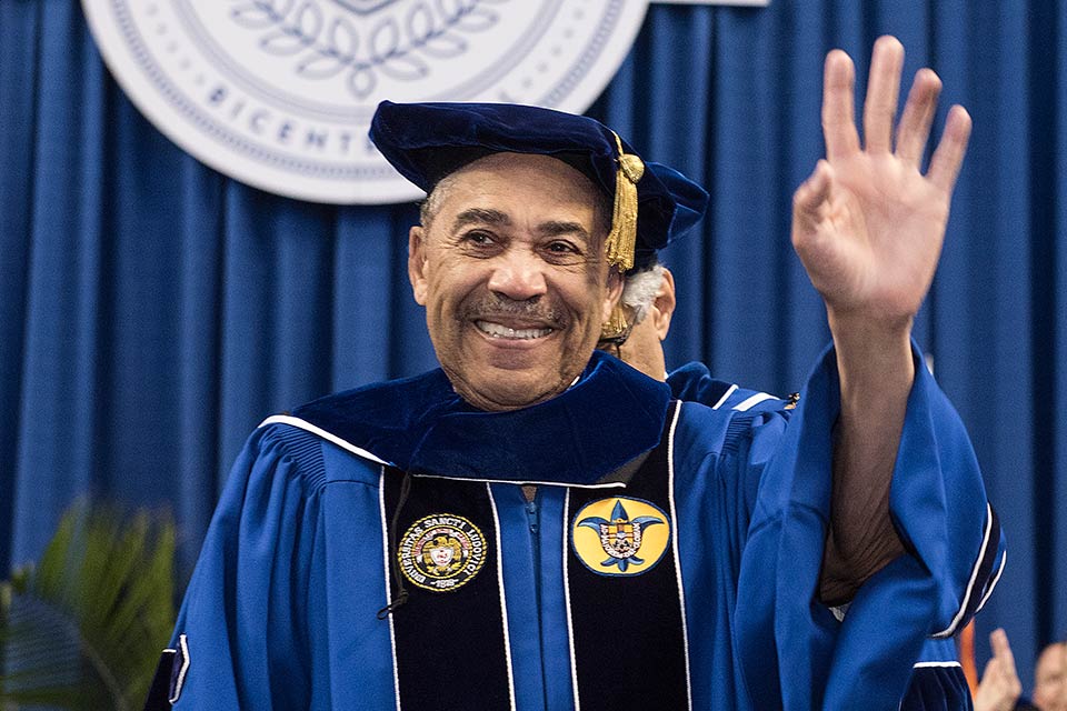 William “Bill” Lacy Clay Sr. smiles and waves to the crowd during the SLU's 2018 commencement ceremony. He wears a blue cap and gown with a gold tassel.