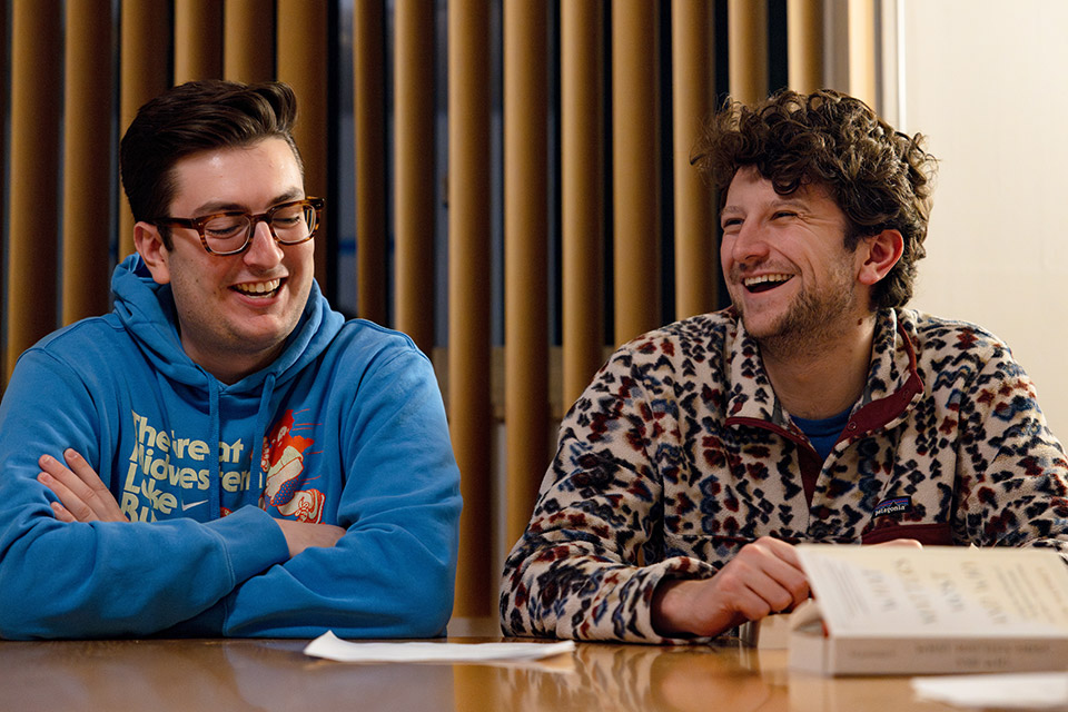 Two male Billiken Teachers sit next to each other at a table, smiling. Two male Billiken Teachers sit next to each other at a table, smiling.