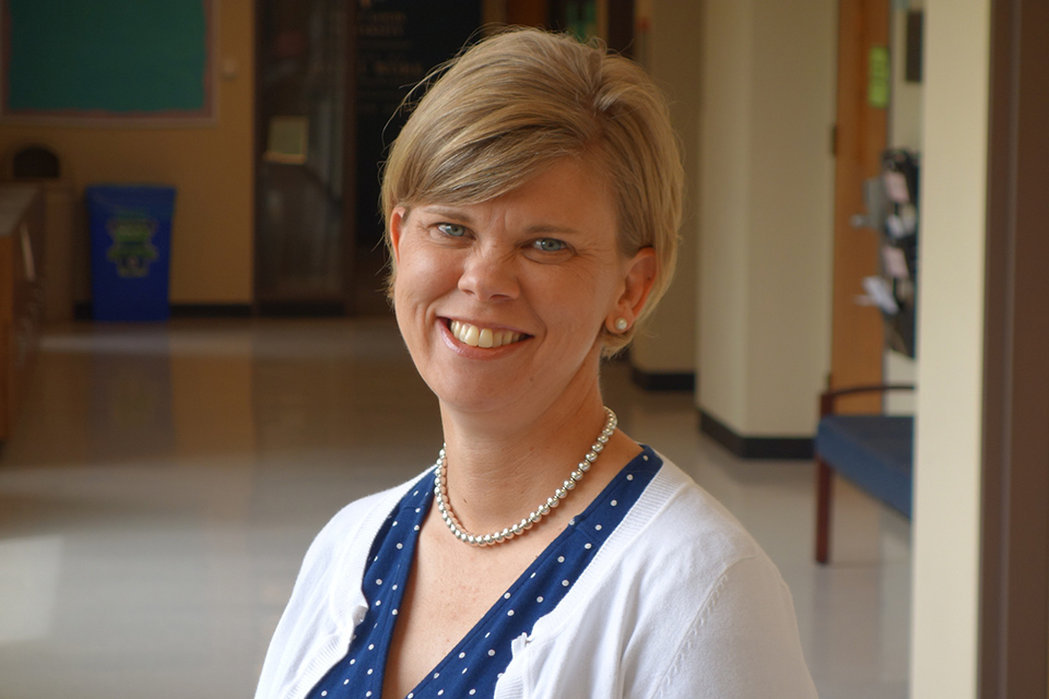 A woman with short blonde hair smiles in a school hallway.