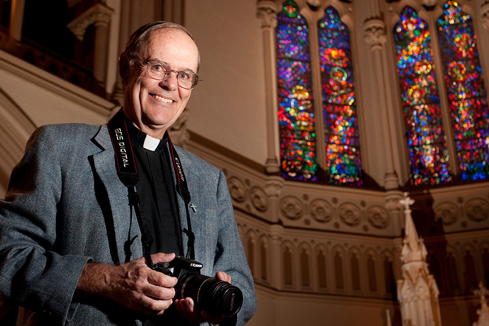 J.J. Mueller, S.J., an older priest wearing glasses, wears a camera around his neck in front of stained glass windows.