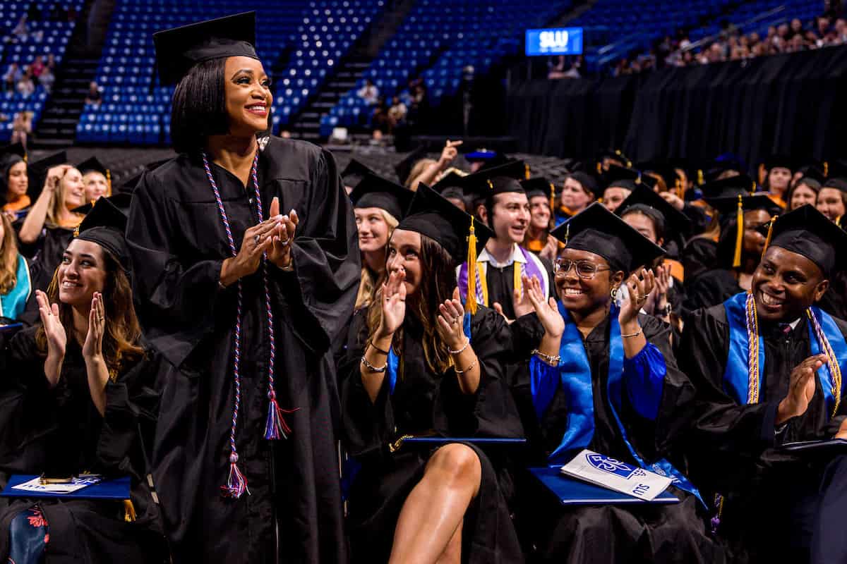 Graduates at commencement wearing caps and gowns. One student stands up, smiling, while others look at her and clap.