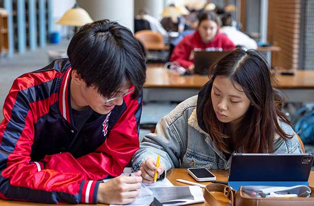A male student and a female student are sitting at a desk in the library, both looking at a piece of paper. A cell phone and an electronic tablet are also on the table.
