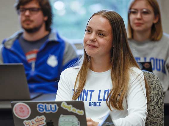 A student wearing a SLU Dance Shirt with a laptop covered in stickers looks attentively toward the front of a classroom