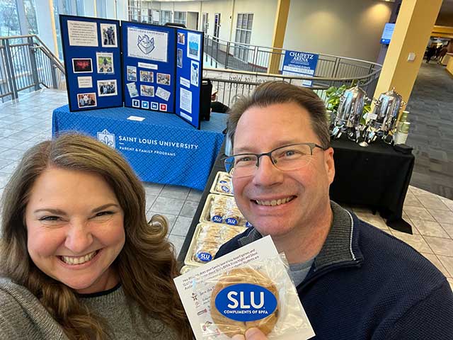 Two SLU parents standing in front of a table showcasing a trifold display of BPFA activities, urns of hot cocoa and cookies for students.