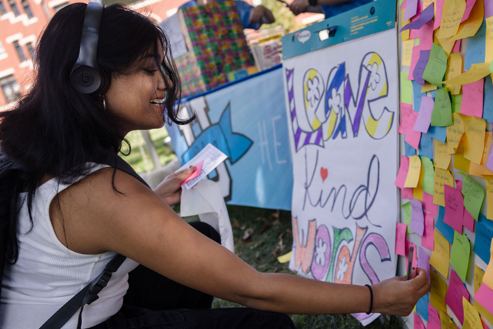 A student wearing headphones examines a wall of sticky notes next to a sign that reads Leave Kind Words.