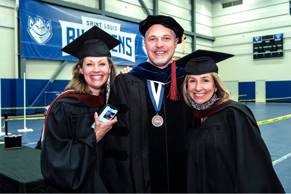 Three professors wearing academic regalia smile while standing in a gymnasium.