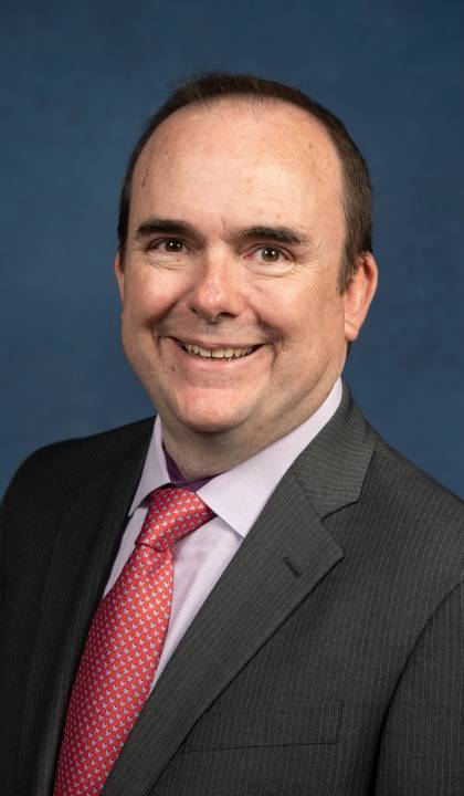 Headshot of Joe Lyons dark suit, light shirt and red tie.