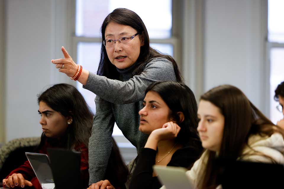 An instructor stands behind a student at a table desk and gestures