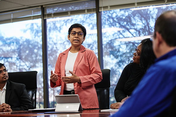 A person wearing professional dress stands next to a conference table while talking to others in a meeting.
