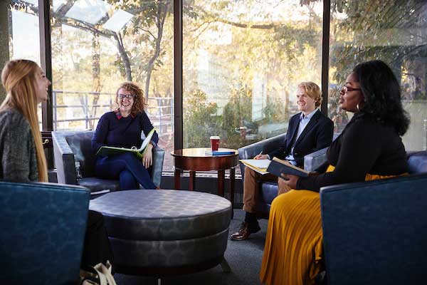Four students sitting in an atrium in navy blue chairs and wearing business casual attire laugh and listen intently to a student telling a story on the left side of the picture.