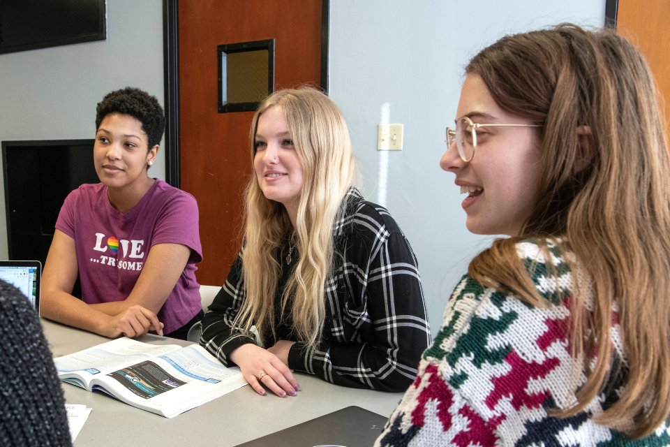 Three female undergraduate students sit at a table around a computer. 