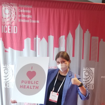 Hannah Buncher, wearing a blue blazer and face mask, stands in front of a conference banner holding a large "I love public health" sign. 