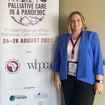 Lydia Thomas, wearing a blue blazer and black pants, stands in front of a banner for a palliative care conference