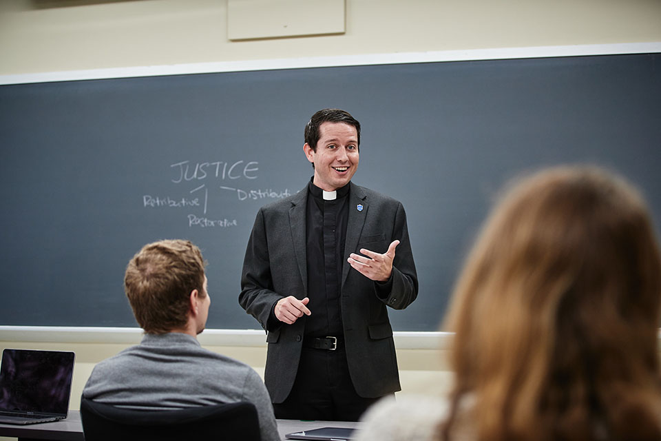 Michael Rozier, S.J. Ph.D., stands in front of a chalkboard speaking to a room full of students about the topic of justice. 