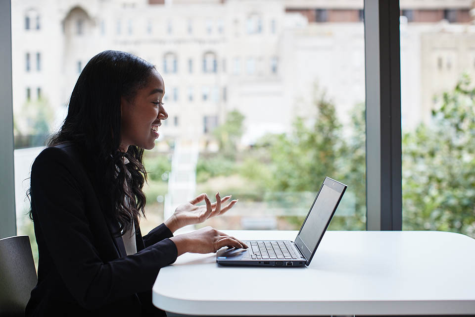 Female student in a suit, sits next to a large window with her computer on a video call. 